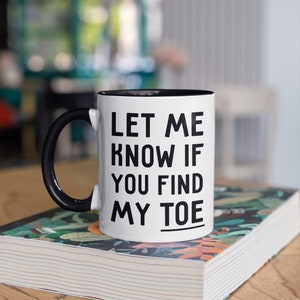 May include: White coffee mug with a black handle and black rim. The mug has black text that reads "Let me know if you find my toe". The mug is sitting on top of a closed book with a colorful floral design.