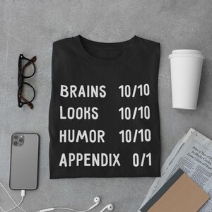 May include: Black t-shirt with white text that reads "BRAINS 10/10 LOOKS 10/10 HUMOR 10/10 APPENDIX 0/1"