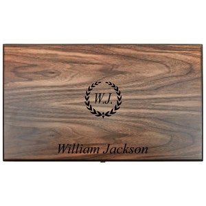 Pode incluir: Uma caixa de madeira retangular com acabamento castanho escuro. A parte superior da caixa apresenta o nome "William Jackson" em uma fonte serifada, abaixo de um emblema circular com as iniciais "W.J." dentro de uma coroa de louros.