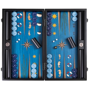 May include: A nautical-themed backgammon set with a blue playing surface. The set features light blue and navy blue checkers, dice, and black dice cups. Decorative elements include seahorses, anchors, and compass rose designs. The set is enclosed in a black case.