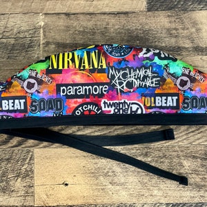 May include: A colorful scrub cap featuring a collage of band names including Nirvana, My Chemical Romance, Paramore, and SOAD. The cap has a black band and ties for a secure fit. The design incorporates various colors and fonts.