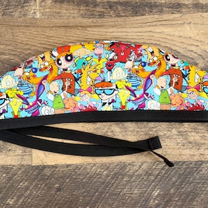 May include: A colorful scrub cap featuring cartoon characters from popular 90s animated shows. The cap has a black band and ties. The design includes characters like Dexter, Doug, and the Powerpuff Girls, set against a light blue background.