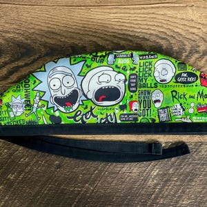 May include: A green and black scrub cap featuring cartoon characters and text from the animated series "Rick and Morty." The cap has a black band and ties. The design includes various characters and phrases.