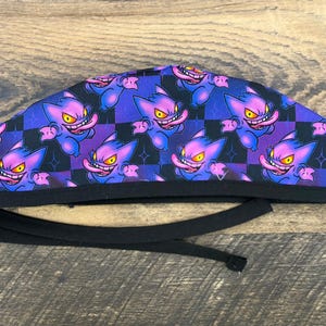 May include: A patterned scrub cap featuring a repeating cartoon character design in purple, blue, and pink. The cap has a black band and ties. The background is a checkerboard pattern.