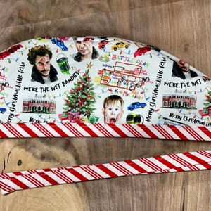 May include: White scrub hat with a red and white candy cane trim. The hat features a repeating pattern of illustrations from the movie Home Alone, including Kevin McCallister, the Wet Bandits, and the iconic Christmas tree. The text "Merry Christmas, little fella" is repeated throughout the pattern.