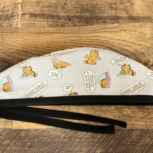 May include: A scrub cap featuring a light gray fabric with a repeating pattern of the orange cartoon cat Garfield, including various speech bubbles with text, finished with a solid black fabric band and black tie straps at the bottom edge.
