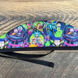 May include: A colorful surgical scrub cap featuring a vibrant design with characters from a popular animated series. The cap has a black band and ties. The design includes characters in a spaceship and other scenes.