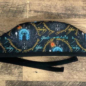 May include: Black scrub cap featuring a repeating pattern of Elvish script and imagery from a fantasy world. The cap includes a black tie for securing. The design includes the text "The One Ring".