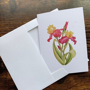 May include: A white greeting card with a watercolor painting of yellow and red iris flowers, with green leaves and stems. The card is inside a white envelope, with the envelope partially open. The background is a dark wood surface.