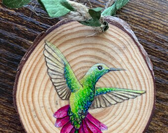 May include: A wooden slice ornament featuring a vibrant hummingbird illustration. The bird has green and blue feathers, with a bright pink tail. The ornament is suspended by a natural rope and green fabric.