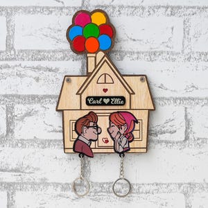Può includere: Un portachiavi da parete in legno a forma di casa con un gruppo di palloncini colorati sulla parte superiore. Al centro è riportato il testo Carl e Ellie con un'icona a forma di cuore. Due sagome di personaggi pendono sotto, fissate a catenelle.