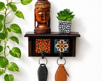 May include: A dark brown wooden wall-mounted key holder featuring two decorative square tiles with orange and blue floral patterns. The unit includes two black metal hooks, each holding a key fob, with a carved wooden Buddha head and a small green succulent in a grey textured pot placed on the top shelf.