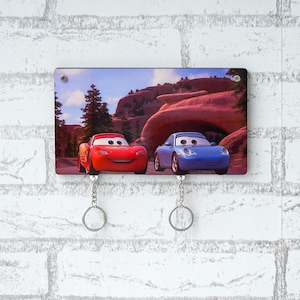 May include: Key holder featuring Lightning McQueen and Sally Carrera from the animated movie Cars. The rectangular holder has a scenic background and two keychains. The key holder is mounted on a white brick wall.