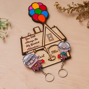 May include: A wooden house-shaped wall plaque featuring a cluster of colourful balloons at the top and the text "You Will Always Be My Greatest Adventure...". Two character keychains labelled "Carl" and "Ellie" are attached to the wooden base.