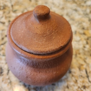 May include: A small, terracotta pot with a lid. The pot is a reddish-brown color and has a smooth, slightly textured surface.