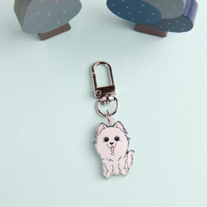 Puede incluir: Un llavero plateado con un dije de perro de dibujos animados blanco. El perro tiene ojos negros y una lengua rosa que sobresale.