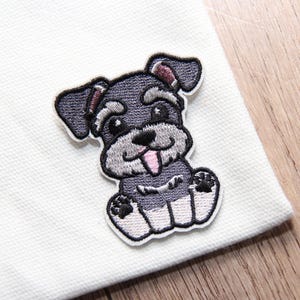 May include: Embroidered patch featuring a cartoon Schnauzer dog in shades of gray, black, and white. The dog has a pink tongue and is sitting with its paws forward. The patch is on a white fabric.