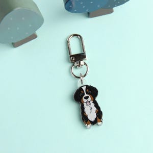 May include: A silver keychain with a black, white, and brown Bernese Mountain Dog charm. The dog is sitting and has a happy expression.