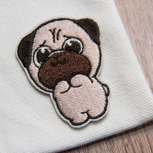 Puede incluir: Parche bordado con un perro pug de dibujos animados con una cara marrón claro, orejas marrones oscuras y una nariz marrón oscura. El pug está bordado con un contorno blanco y está sobre un fondo de tela blanca. La expresión del pug es triste.