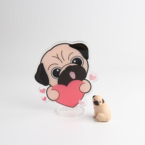 Op de afbeelding: Een transparant acryl beeldje van een cartoon-mops die een roze hart vasthoudt. De mops heeft zwarte ogen en een zwarte neus. Er zijn kleine roze hartjes rond de mops. Een klein, beige mopsbeeldje zit op een wit oppervlak.