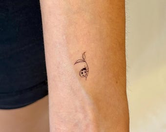 Puede incluir: Un pequeño tatuaje de tinta negra de líneas finas en el antebrazo que presenta una mariquita minimalista posada sobre una planta delicada de tallo fino con dos hojas pequeñas. El diseño utiliza líneas simples y limpias para el insecto.