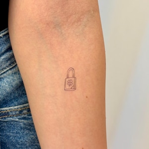 May include: A small, fine-line black ink tattoo on skin depicting a rectangular padlock with a rounded shackle. The center of the padlock features a simple, four-leaf clover outline design, rendered in a minimalist, clean, and precise style.