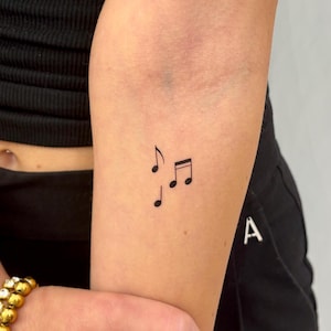 Op de afbeelding: Een minimalistische tatoeage van drie zwarte muzieknoten op een arm. De noten zijn een G-sleutel en twee achtste noten, wat een eenvoudig maar elegant ontwerp creëert. De tatoeage is op de binnenkant van de onderarm geplaatst.