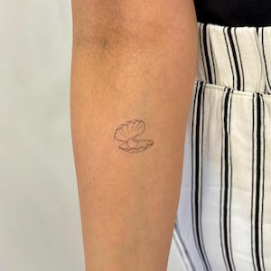 Puede incluir: Un pequeño tatuaje minimalista de una concha abierta con una perla en su interior. El tatuaje es un simple dibujo lineal en tinta negra, ubicado en el brazo. El tono de piel es claro y el fondo está borroso.