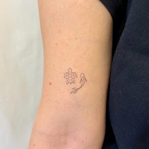 Peut inclure: Un tatouage minimaliste représentant une tortue et un poisson. La tortue est représentée avec une carapace détaillée, et le poisson a des lignes fluides. Le tatouage est sur le bras, avec le teint de la peau visible.