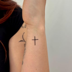 Op de afbeelding: Een kleine, minimalistische kruistattoo op de pols. Het zwarte inktkruis is eenvoudig en elegant, met strakke lijnen. De tattoo bevindt zich aan de binnenkant van de pols en de huidskleur is licht. De achtergrond is een effen witte muur.