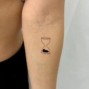 May include: A simple line drawing tattoo of an hourglass with sand in the bottom half.