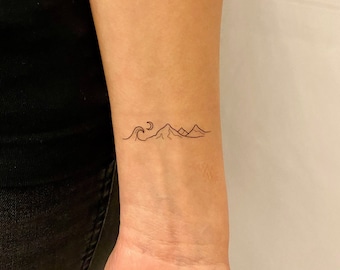 May include: A minimalist tattoo design featuring a wave, crescent moon, and mountain range. The black ink tattoo is on the wrist, showcasing a simple yet elegant landscape scene. The tattoo is a small, delicate design.