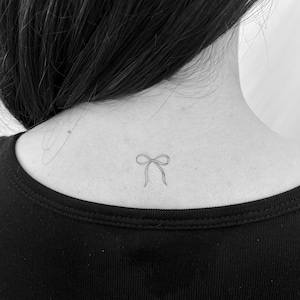 Peut inclure: Un petit tatouage de nœud délicat sur la nuque d'une personne. Le tatouage est un dessin simple et fin à l'encre noire, avec le nœud noué au centre et les extrémités qui coulent vers le bas. La personne porte un haut noir.