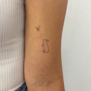 Puede incluir: Un diseño de tatuaje minimalista que presenta un pequeño conejo mirando hacia arriba a una mariposa. El tatuaje de contorno negro está en el brazo de una persona. La mariposa está encima del conejo con unos pequeños puntos en medio.