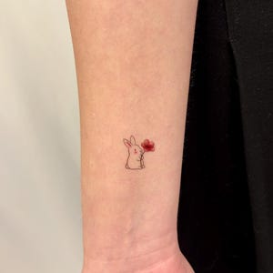 Könnte beinhalten: Minimalistisches Tattoo eines Cartoon-Hasen, der eine rote Blume hält. Das Tattoo befindet sich am Handgelenk. Der Hase ist mit feinen Linien gezeichnet und hält die Blume. Das Design ist einfach und zart.