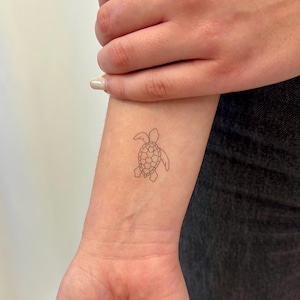 Peut inclure: Un petit tatouage minimaliste d'une tortue de mer sur un poignet. Le tatouage est un simple contour noir, représentant la carapace, la tête et les nageoires de la tortue. La peau est claire, et le fond est neutre.