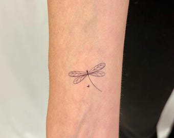 Puede incluir: Un tatuaje de tinta negra con líneas finas que representa una libélula sobre la piel. El diseño presenta líneas delicadas y delgadas que forman las alas del insecto con un patrón de venas detallado y un cuerpo esbelto que termina en una curva.