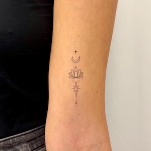 Puede incluir: Un diseño de tatuaje minimalista con una luna creciente, una flor de loto y una estrella. El tatuaje de tinta negra está en el brazo, con los símbolos dispuestos verticalmente. El diseño incluye pequeños puntos y líneas.