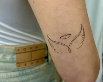 May include: A simple line drawing tattoo of angel wings with a halo above them on a person's arm.