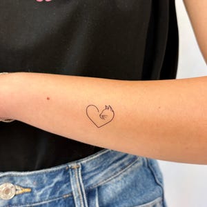 Könnte beinhalten: Ein temporäres Tattoo mit schwarzer Tinte auf der Haut, das eine minimalistische Strichzeichnung eines Katzenprofils in Herzform zeigt. Das Motiv befindet sich auf einem Unterarm, dazu ein schwarzes T-Shirt mit dem rosa Text THE MOOD.