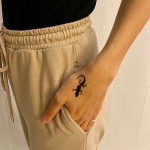 Könnte beinhalten: Eine Hand mit einem temporären Tattoo eines schwarzen Eidechsen.