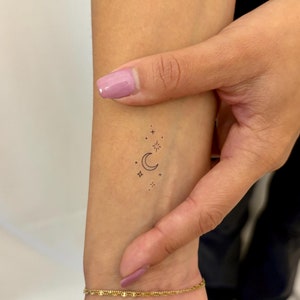 May include: A small, simple tattoo of a crescent moon and stars on a person's wrist.