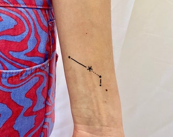 May include: A black line art tattoo on a wrist. The tattoo features a constellation design with a line, a star, and several dots. The person is wearing red and blue patterned pants.
