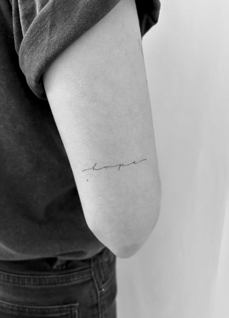 May include: A black and white photo of a person's arm with a simple cursive tattoo that says "hope" on the inside of their elbow.