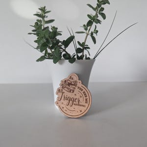 Peut inclure: Un dessous de verre en bois avec les mots "Trigger Warning" et "You Had Me" gravés. Le dessous de verre présente un motif de crâne et de livre. Un vase blanc avec des plantes vertes est en arrière-plan.