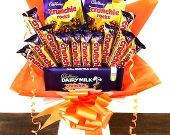 May include: A gift basket filled with Cadbury Crunchie chocolate bars and Crunchie Rocks. The basket is decorated with orange tissue paper and a large orange bow. The basket includes a Cadbury Dairy Milk Crunchie bar.