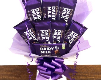 Może przedstawiać: Bukiet tabliczek czekolady Cadbury Dairy Milk owinięty w fioletowy papier tiulowy i przewiązany fioletową wstążką. Bukiet znajduje się w białym pudełku.