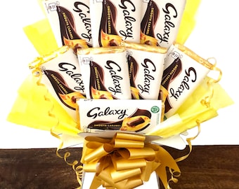 May include: A white gift box filled with Galaxy chocolate bars. The box is decorated with a gold bow and yellow tissue paper. The chocolate bars are wrapped in silver foil and have the Galaxy logo printed on them.