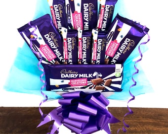 Może przedstawiać: Bukiet tabliczek czekolady Cadbury Dairy Milk ułożonych w przezroczystym pojemniku z fioletowym papierem tiulowym i dużą fioletową kokardką. Tabliczki czekolady są owinięte w fioletową folię z logo Cadbury Dairy Milk i napisem "Marvelous Creations".