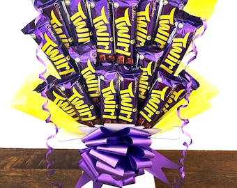 May include: A bouquet of chocolate bars wrapped in purple foil with the word "Twirl" printed on them. The bouquet is arranged in a white box with a purple bow. The box is sitting on a wooden surface.